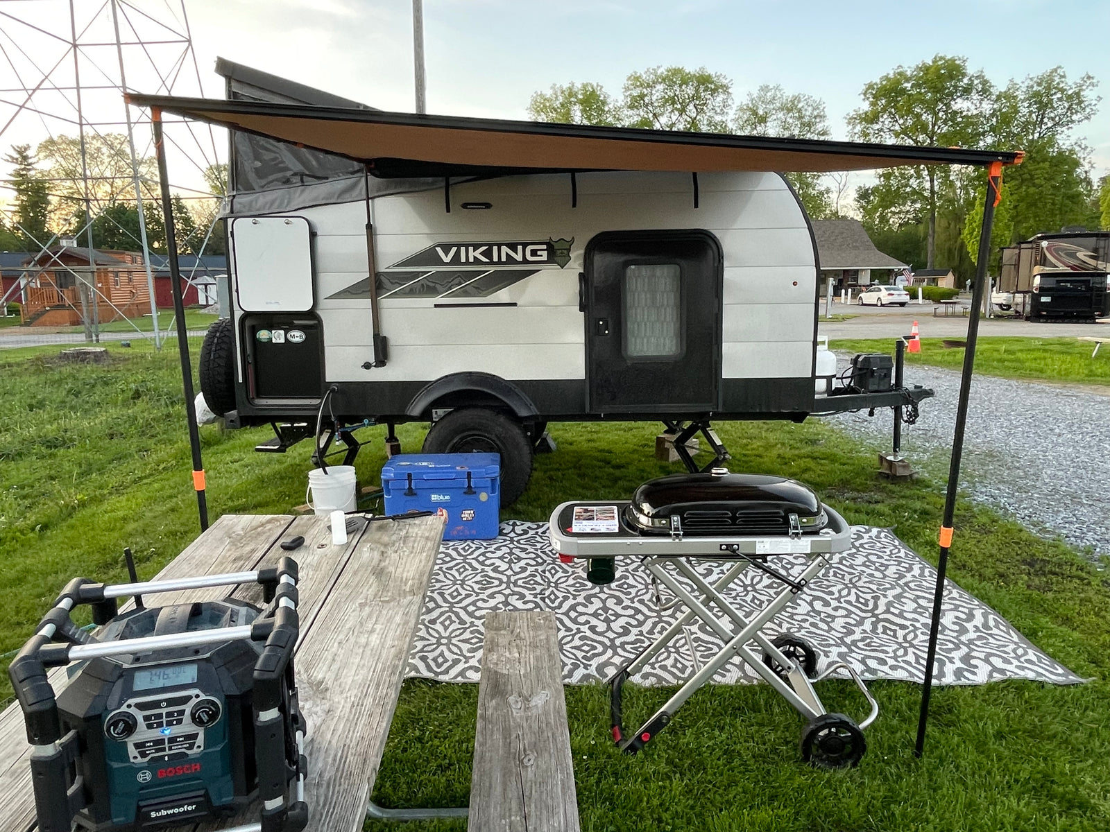 This RV rental showcases a compact Viking travel trailer with an extended awning, outdoor grill, and picnic table at a serene campground.