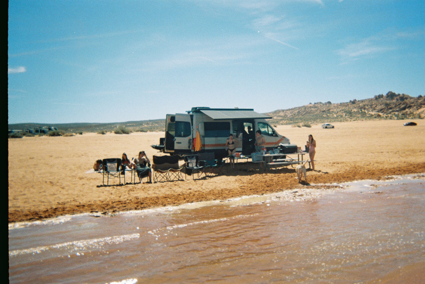 An RV rental offers the perfect setup for friends and a dog enjoying a sunny day of beach camping by the water.