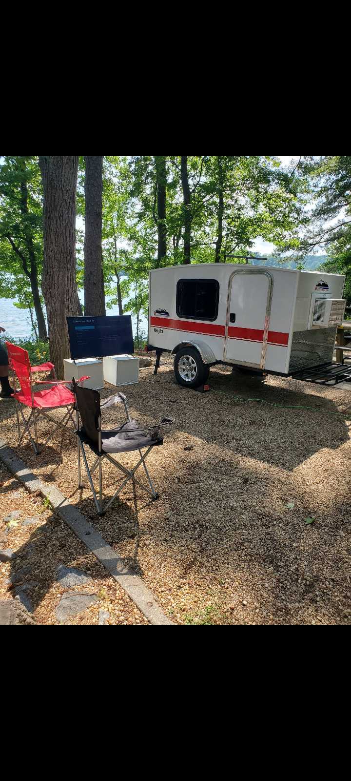 This lakeside RV rental features a white teardrop camper, camping chairs, and an outdoor TV set up on gravel. Enjoy glamping!