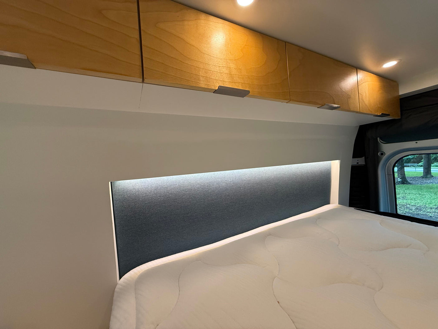 Cozy RV rental interior featuring bright wooden overhead cabinets, an illuminated headboard, and a comfy bed.