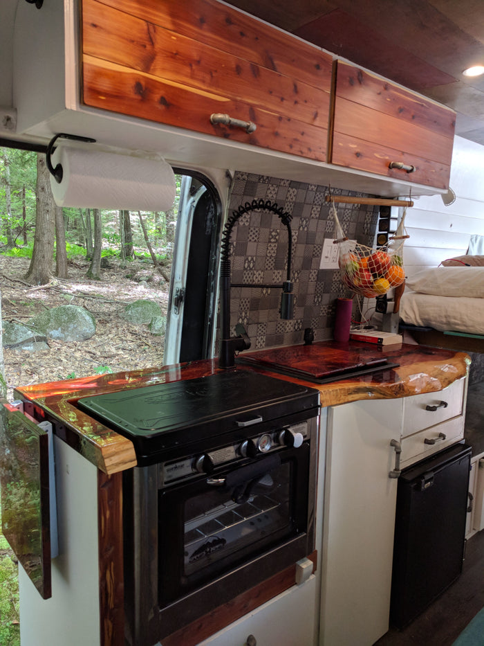 RV rental kitchen area: custom wood counters, oven, sink, and forest view from a camper van.