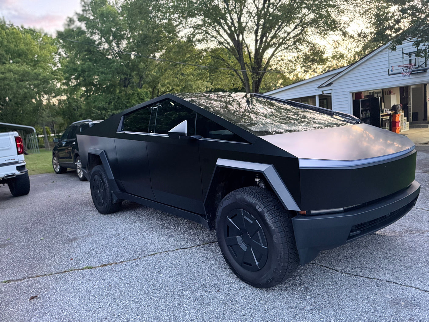A matte black Tesla Cybertruck with a white pickup & SUV on a driveway. Great for a road trip, or an RV rental.
