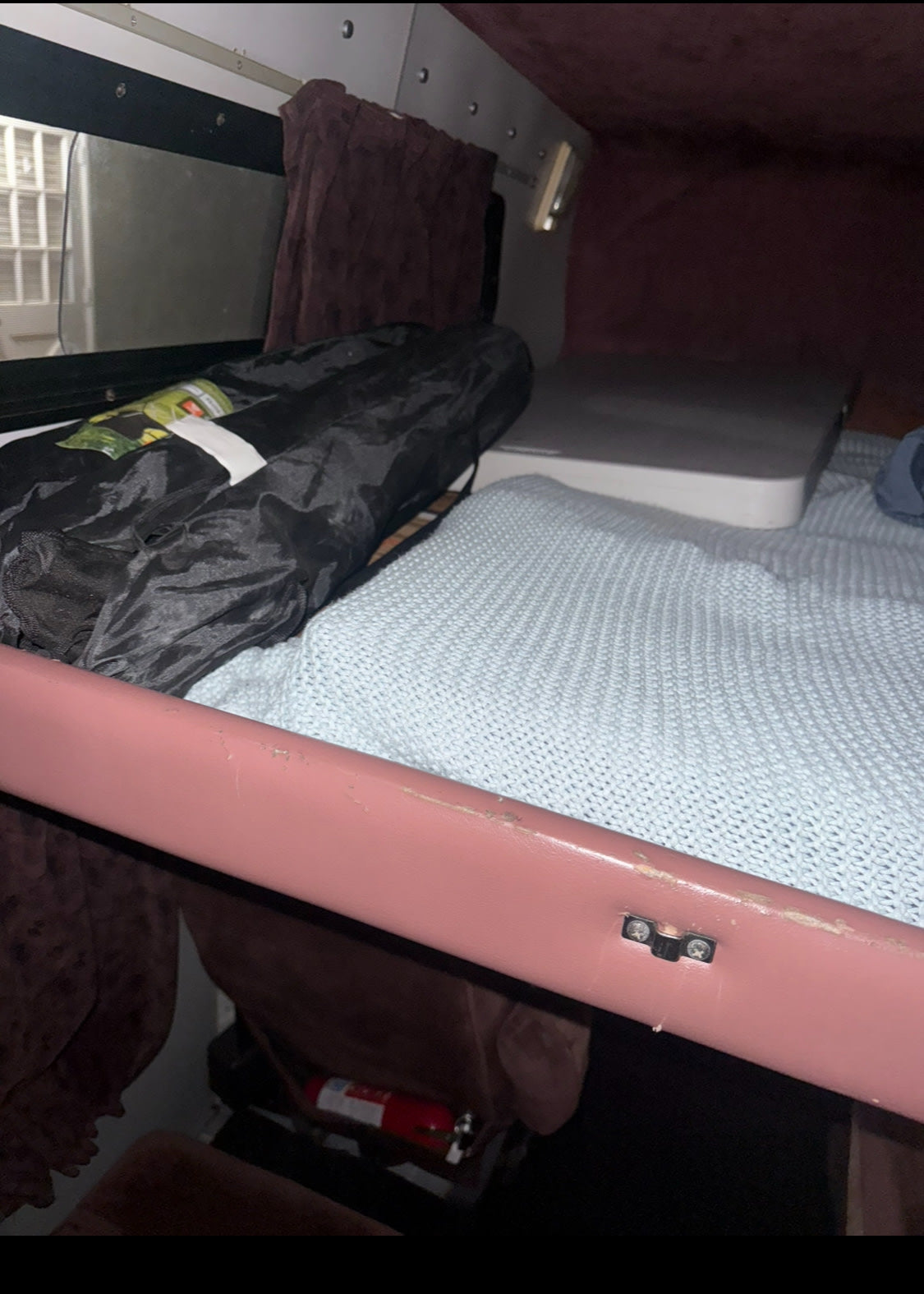 RV rental bunk bed with a light blue blanket, rolled black bag, window, and fire extinguisher below. Ready for adventure.