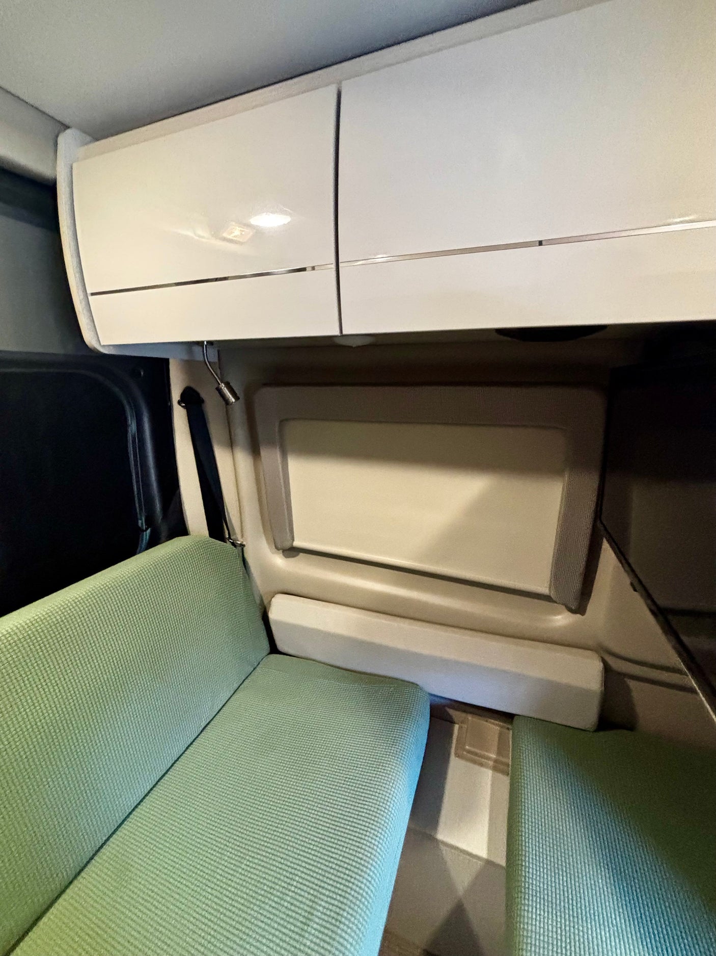 Cozy RV rental interior featuring light green ribbed seating, white overhead storage cabinets, and a window with a shade.