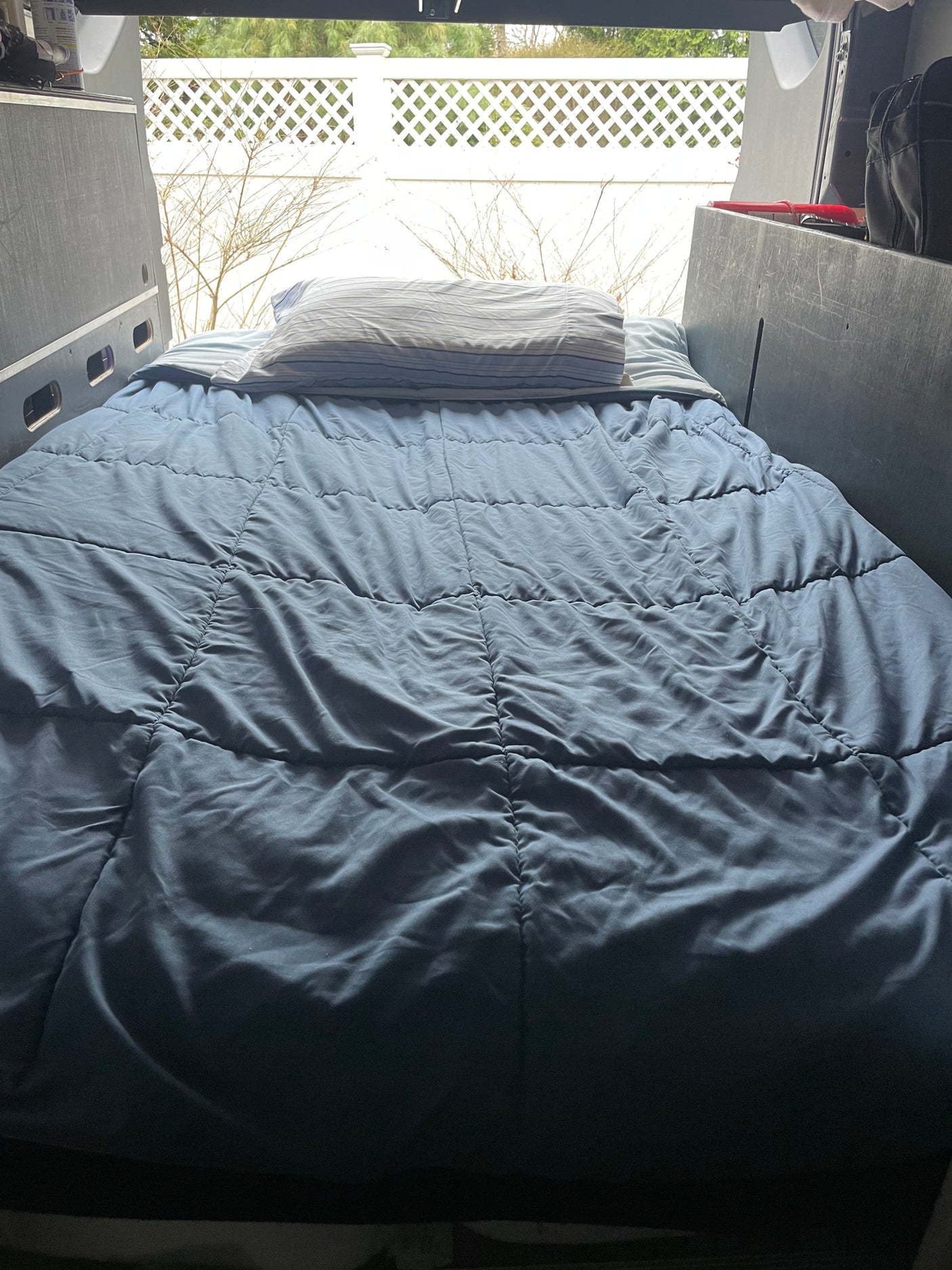 Cozy bed inside an RV rental, offering a view of a white fence and bare trees. Ideal for road trips.