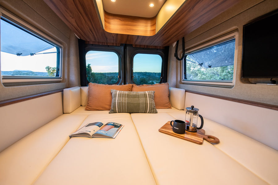 Cozy RV rental interior: lounge bed with coffee, magazine, and scenic views from multiple windows. Perfect for van life adventures!