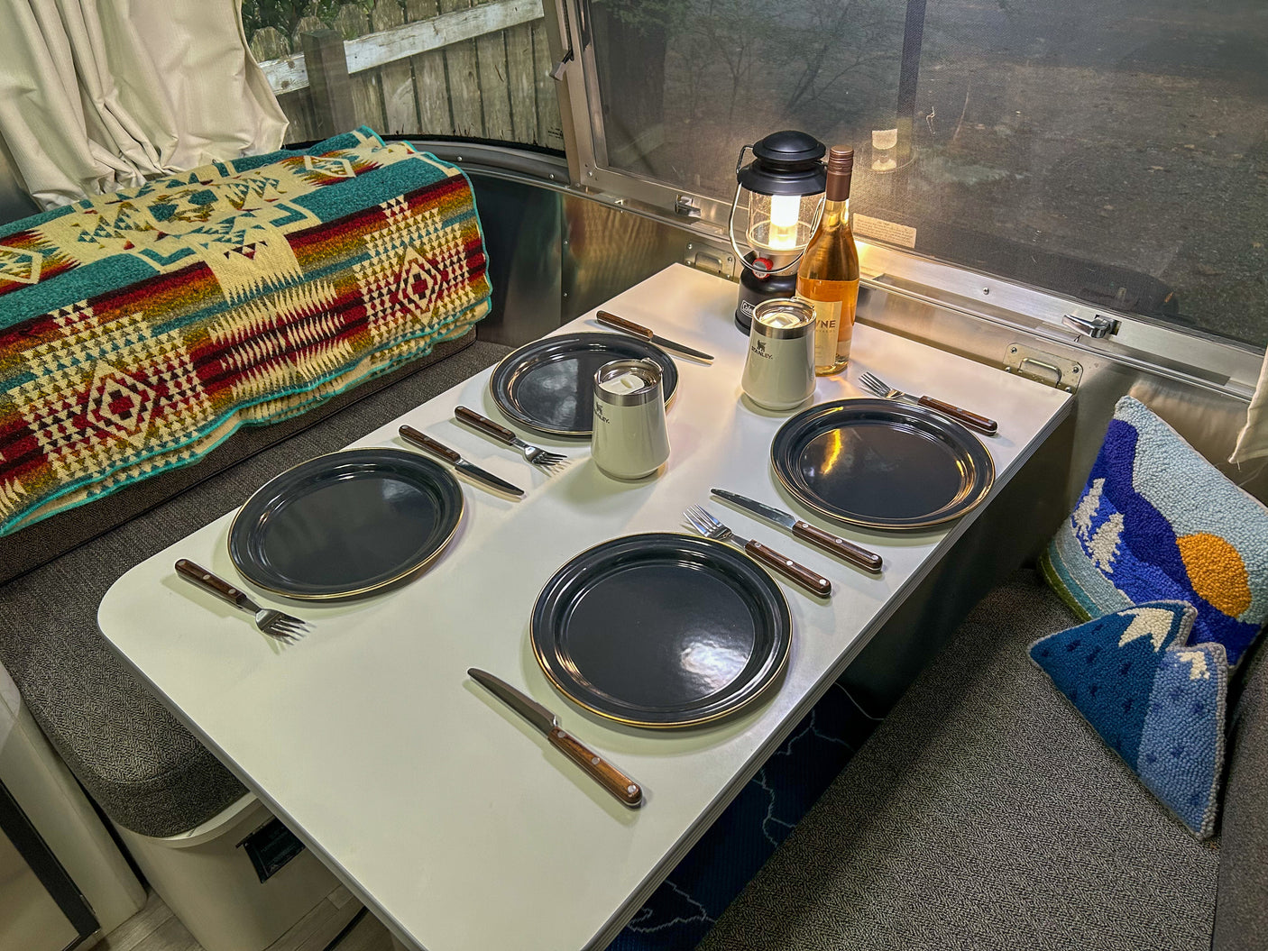 A welcoming RV rental interior features a dining table set for four with dark plates, along with colorful blankets and pillows.