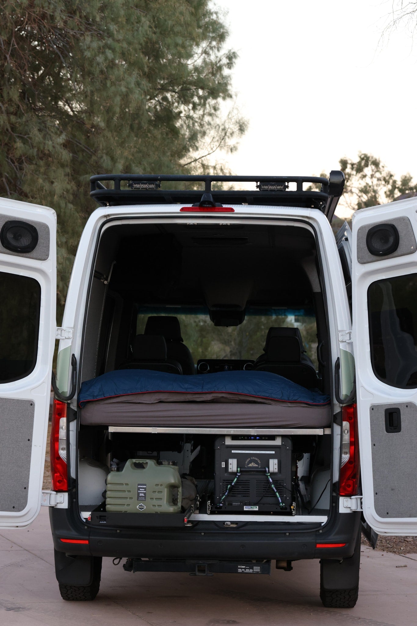 Explore this spacious RV rental camper van with open rear doors, revealing a comfortable bed, portable fridge, and water storage. — photo 11
