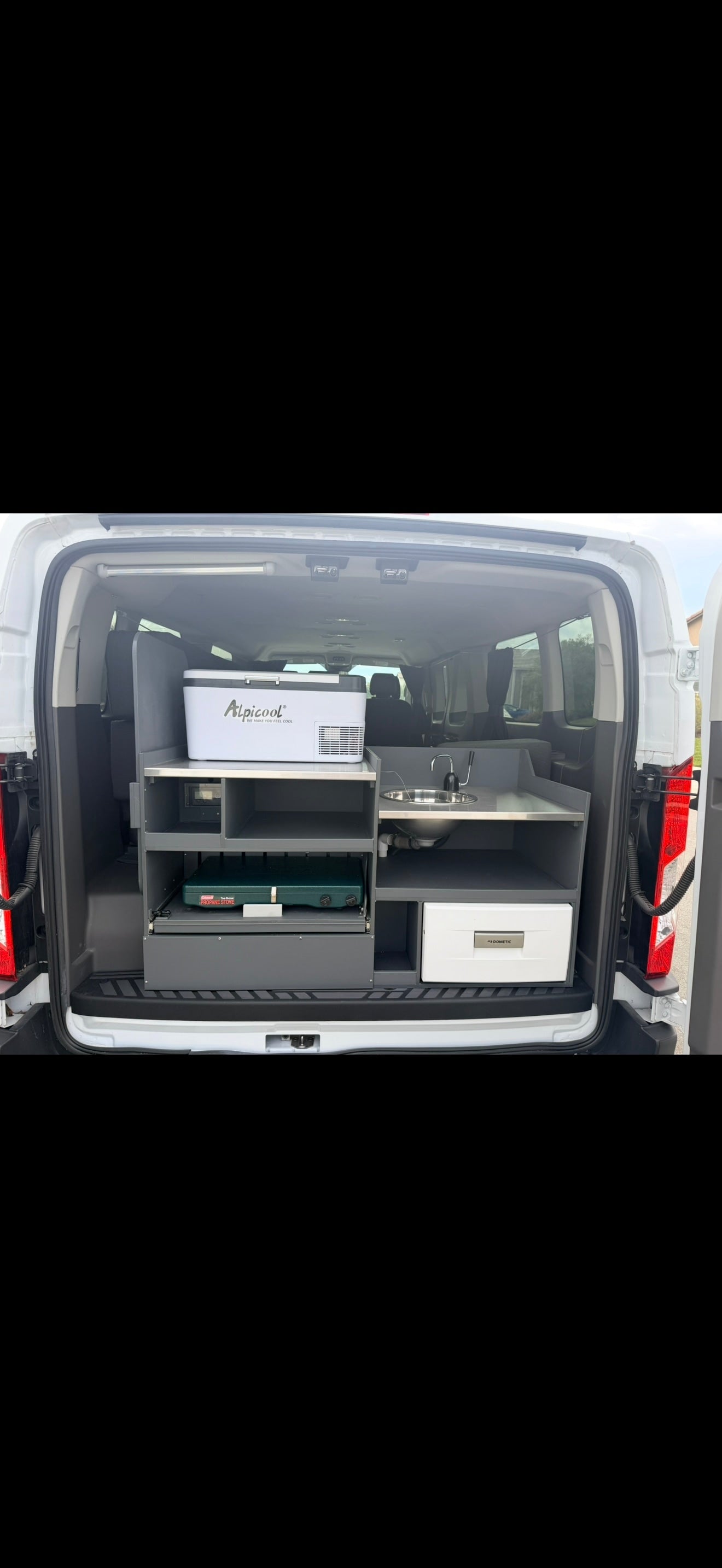 This RV rental shows a compact kitchen setup in a van, featuring an Alpicool cooler, sink, Coleman stove, & Dometic fridge.