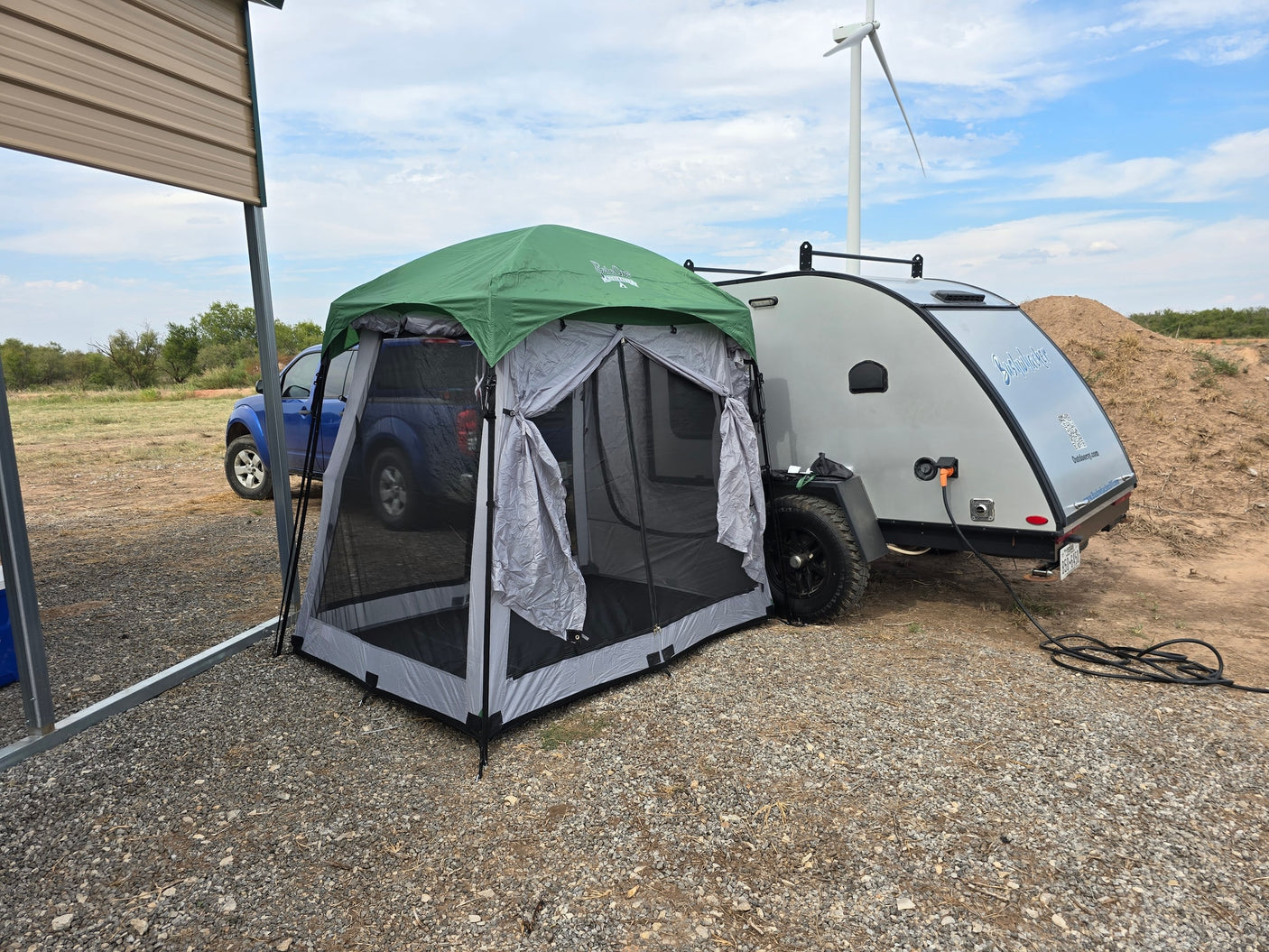 RV rental setup with a teardrop camper, screen tent, and truck on a gravel lot near a wind turbine.