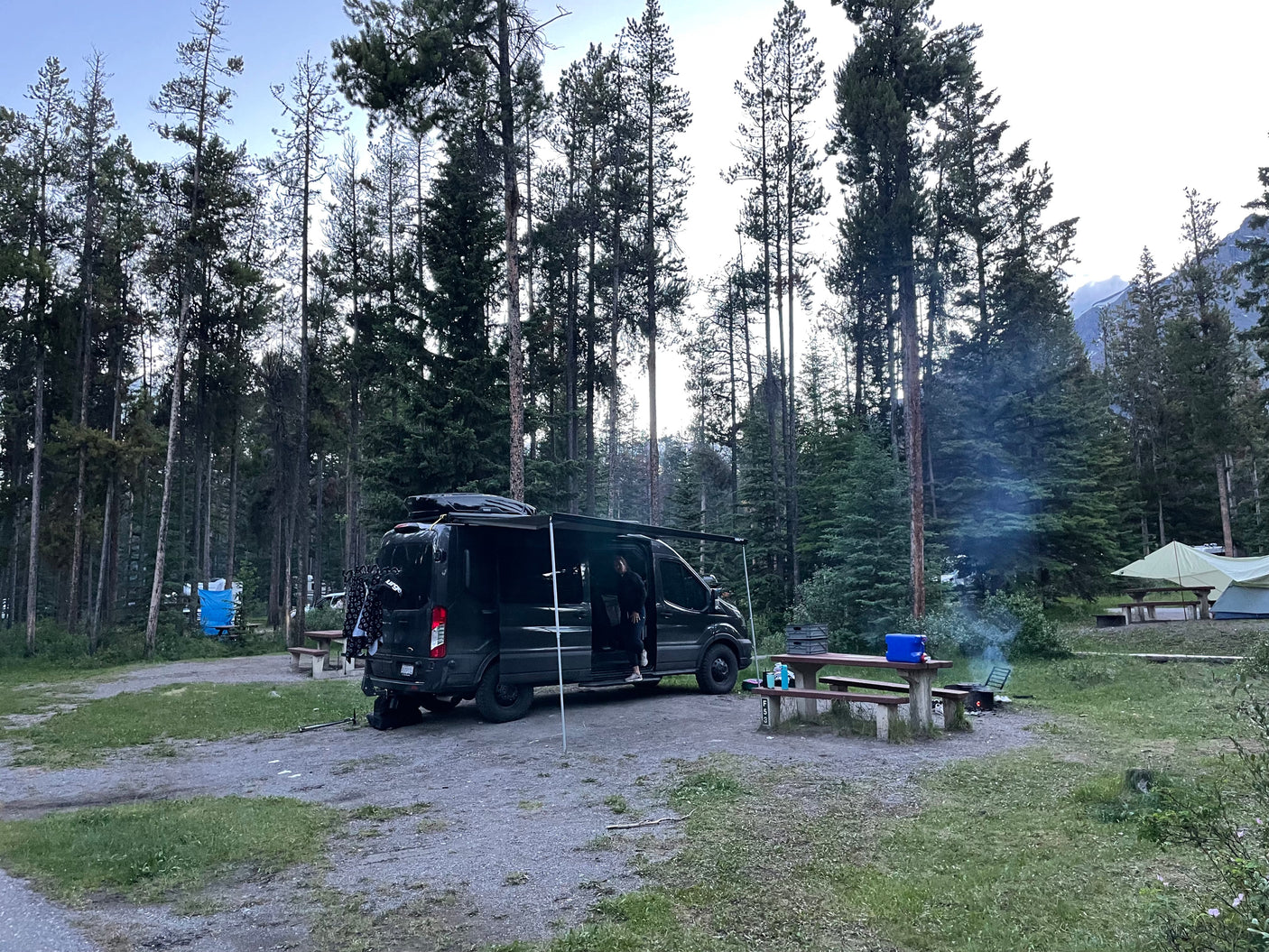 Dark RV rental camper van with awning at a forest campsite. A campfire smokes near a picnic table amidst tall trees.