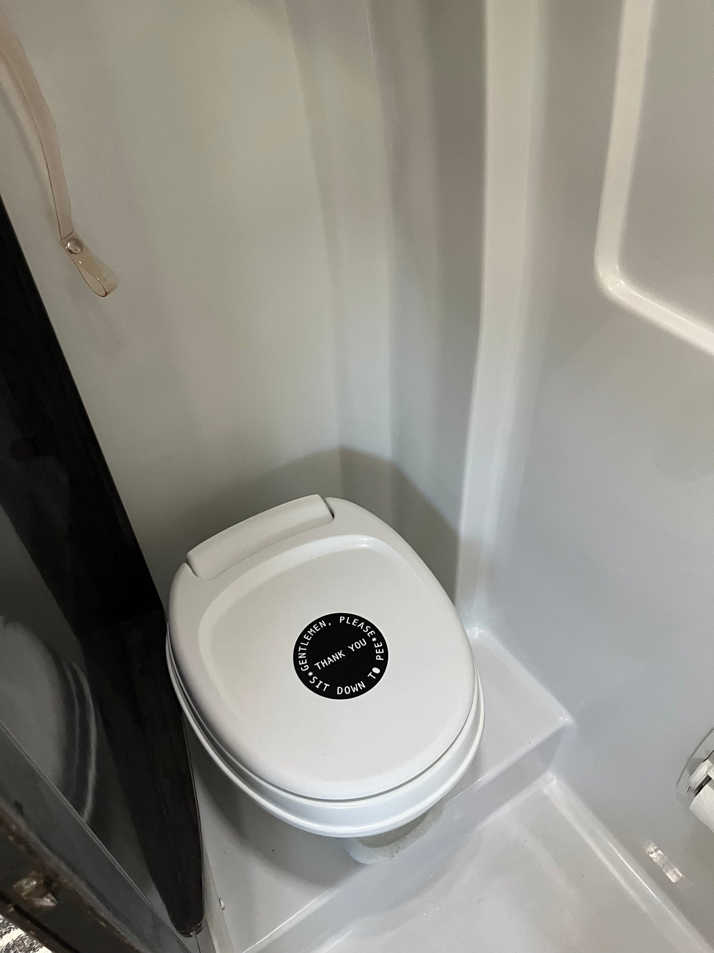 RV rental bathroom features a compact white toilet with a lid sign: 'GENTLEMEN, PLEASE PEE, THANK YOU, SIT DOWN TO'.