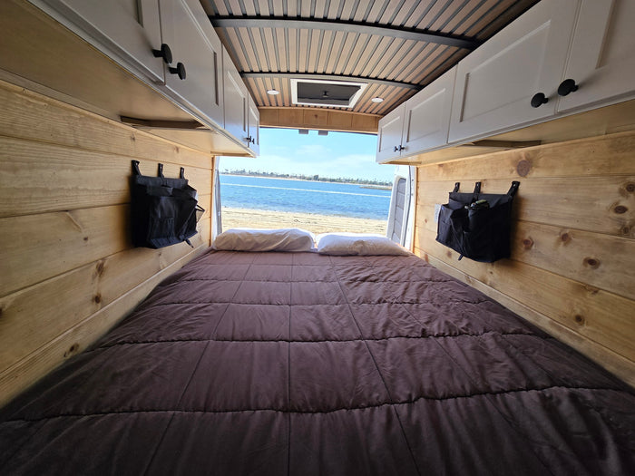 parked on a sandy shore overlooking a calm bay on a sunny day.|RV rental: Rear view of a camper van's wood-paneled interior