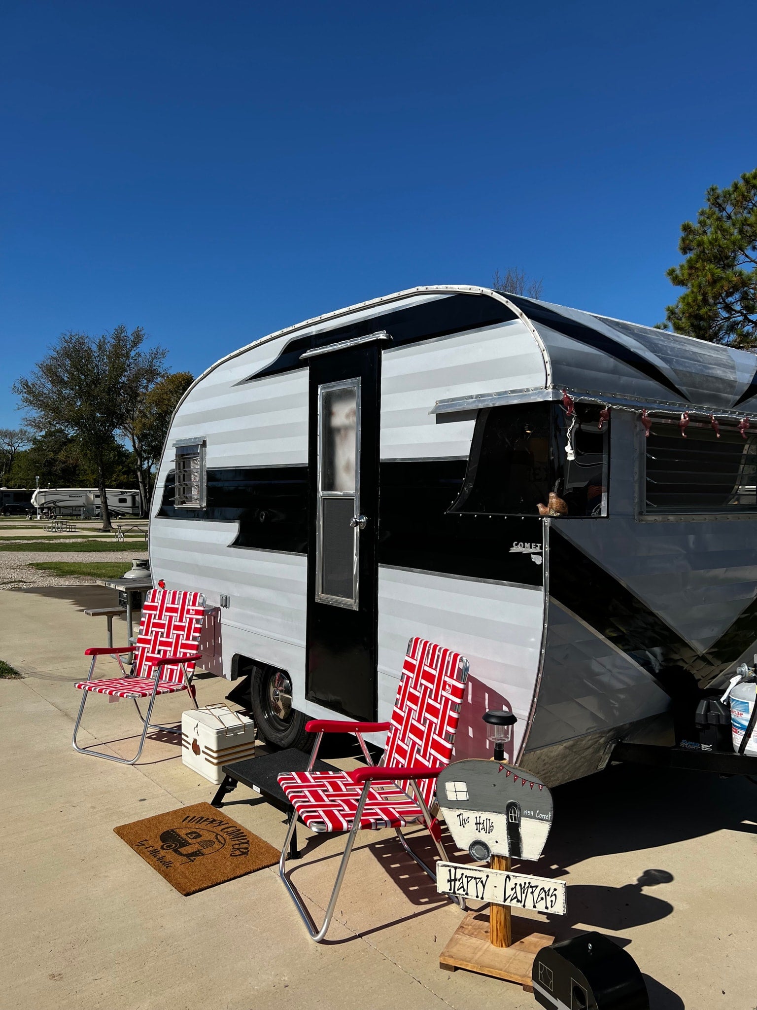 Classic RV rental setup: A black and white vintage travel trailer with red chairs, welcome mat, and decor at a sunny campsite.