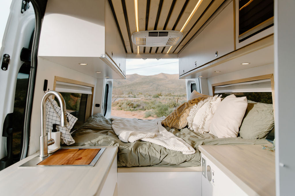 Inside a spacious RV rental: cozy bed, sink, and cabinets, with a desert landscape visible through the rear doors. Ideal for van life.