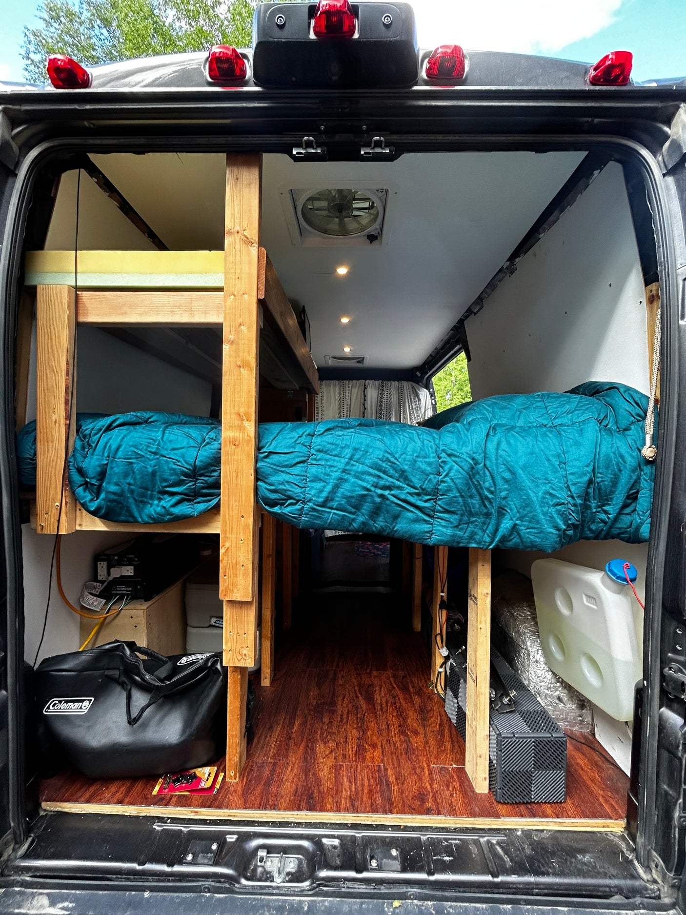 Interior view of an RV rental camper van from the back, revealing custom wooden bunk beds, a single bed, and amenities.