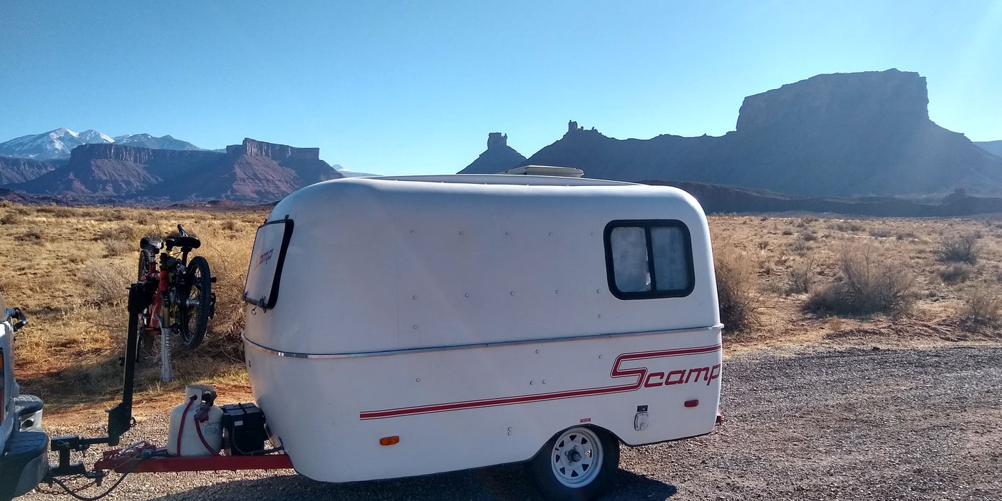 This rv rental shows a compact white Scamp trailer with a bicycle attached, set against a wide desert landscape and snow-capped mountains.