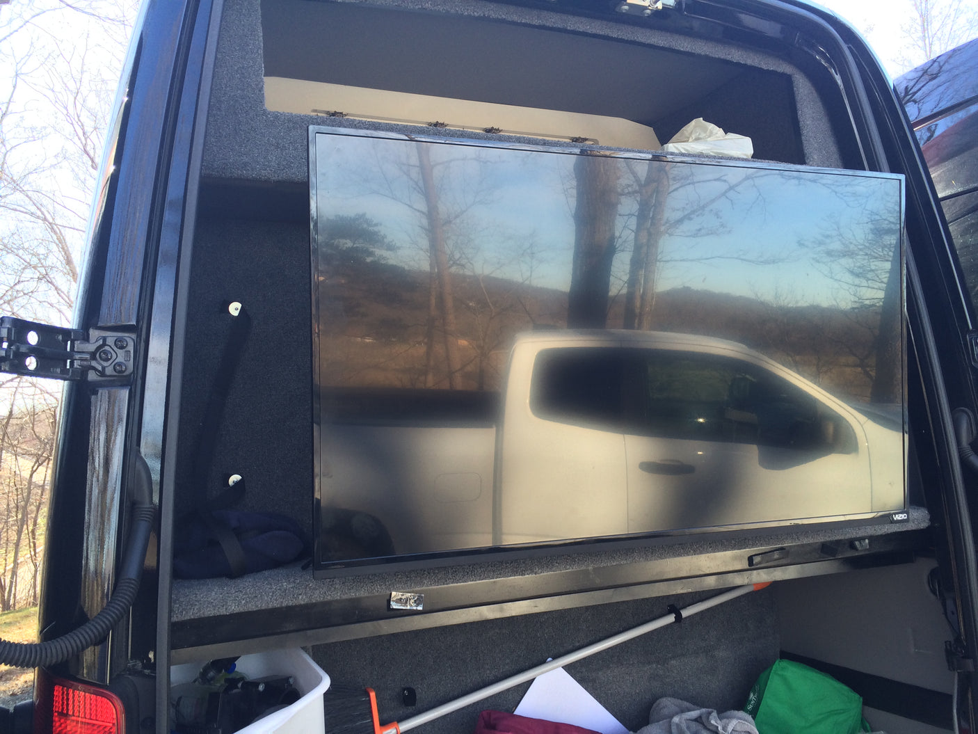 Customized RV rental interior with a large TV and storage. Reflections of a truck & trees are visible on the screen.