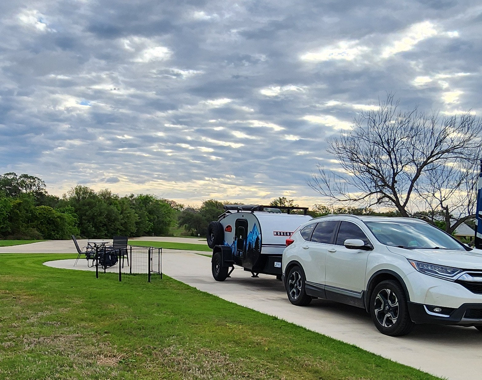 parked at an RV site. Ideal for an RV rental outdoor adventure.|White SUV towing a sleek RV rental teardrop trailer in a paved campsite with a patio and grass