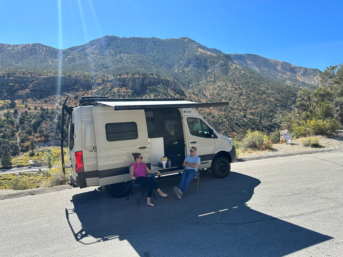 Relaxing outside an **rv rental** with mountain views. A couple enjoys the shade of an awning by their camper van.