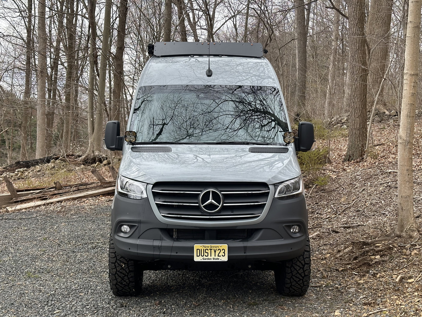 Grey Mercedes Sprinter rv rental van with off-road tires, parked on a gravel path in a forest.