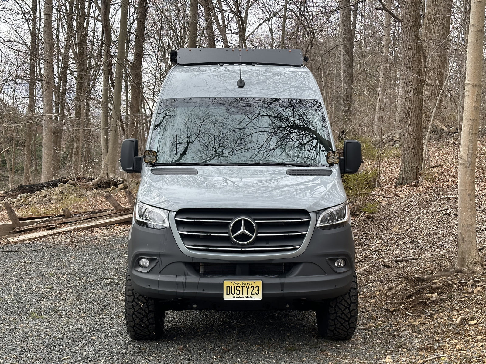 Grey Mercedes Sprinter rv rental van with off-road tires, parked on a gravel path in a forest.