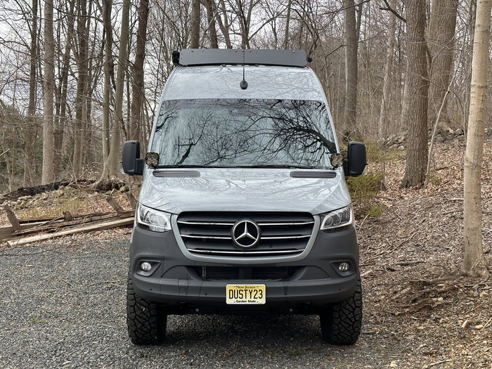 Grey Mercedes Sprinter rv rental van with off-road tires, parked on a gravel path in a forest.