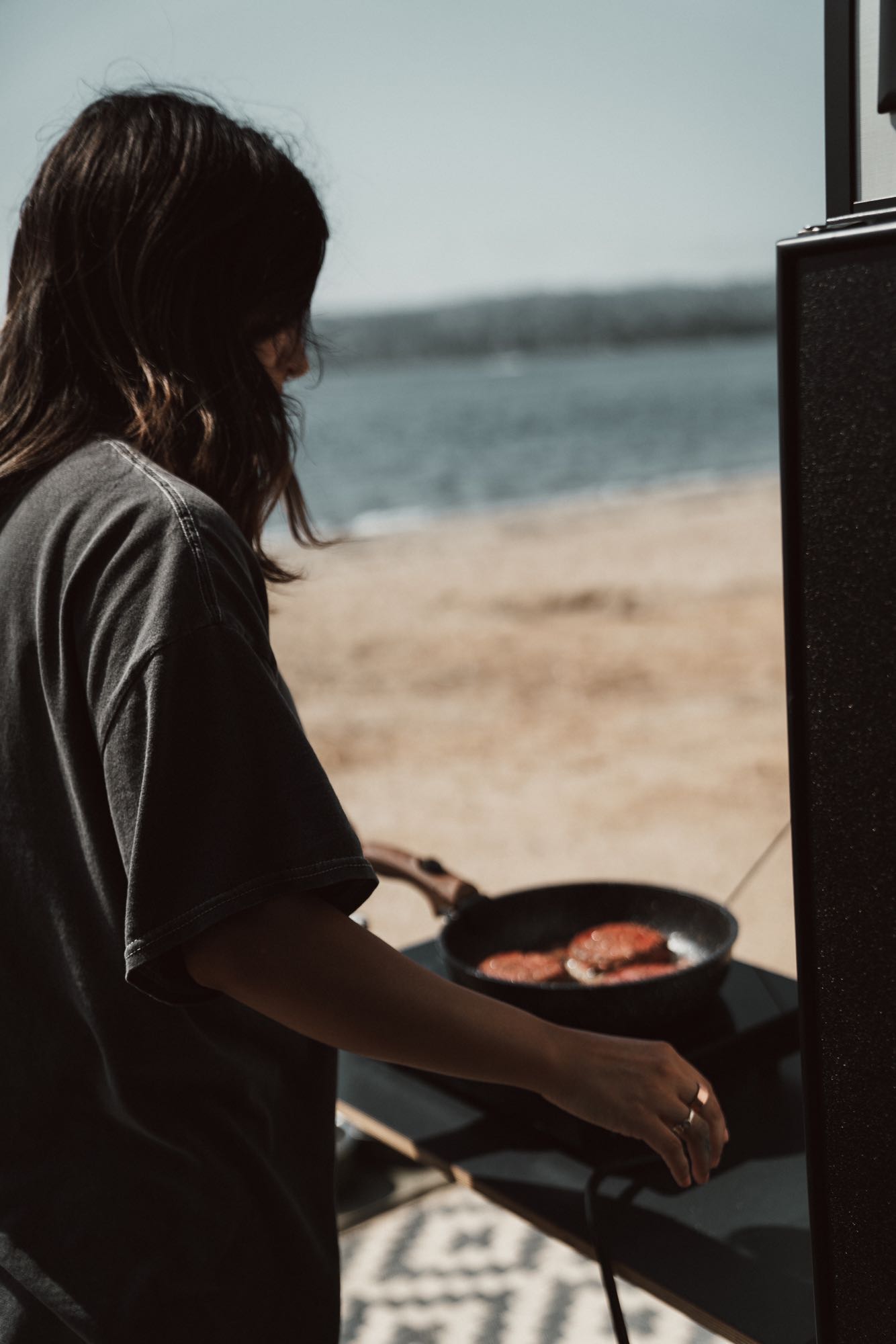 A traveler cooks breakfast patties on a portable stove by the water during an RV rental adventure.