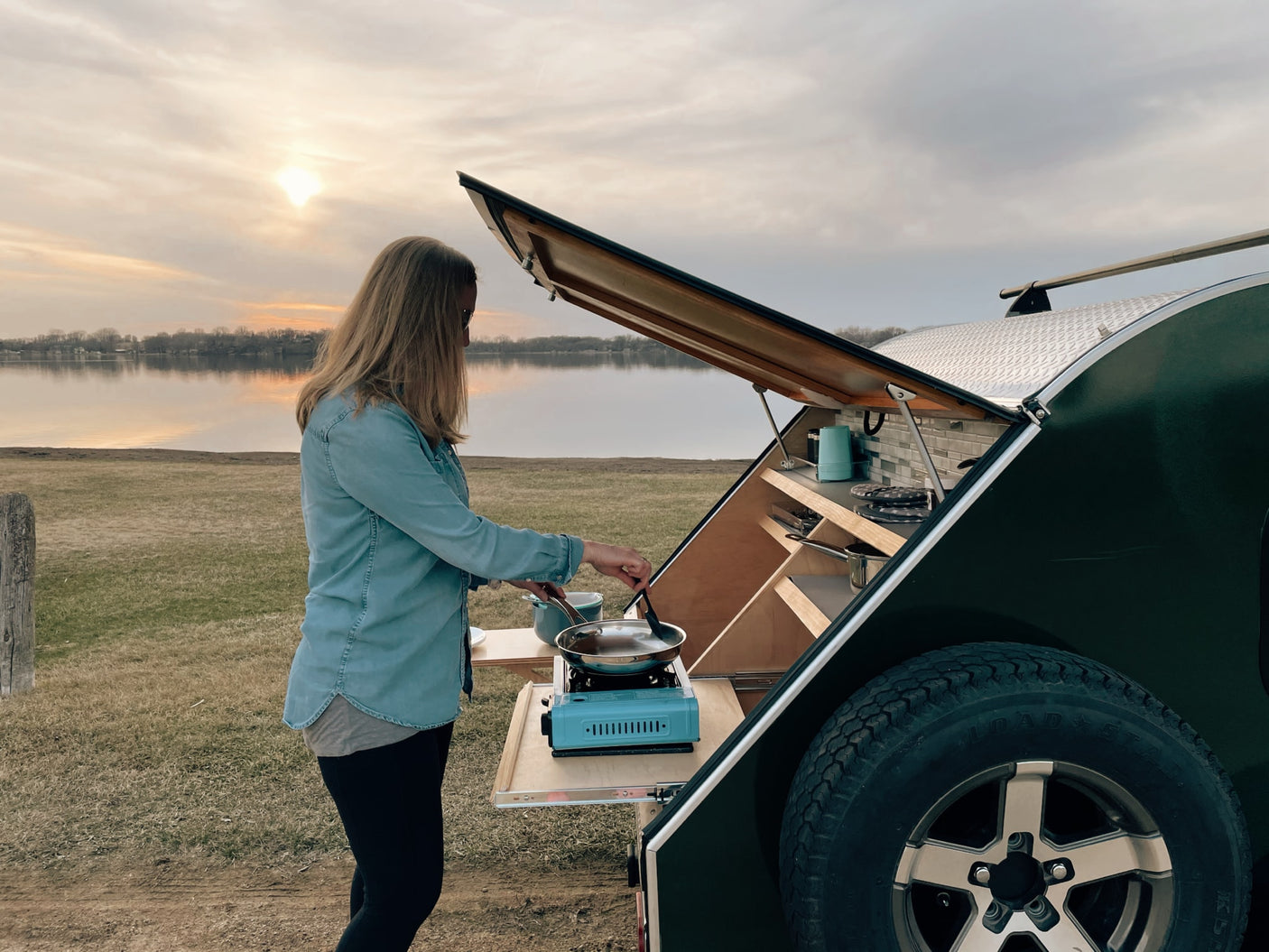 RV rental adventure: Woman cooking lakeside from her teardrop trailer's compact outdoor kitchen at sunset.