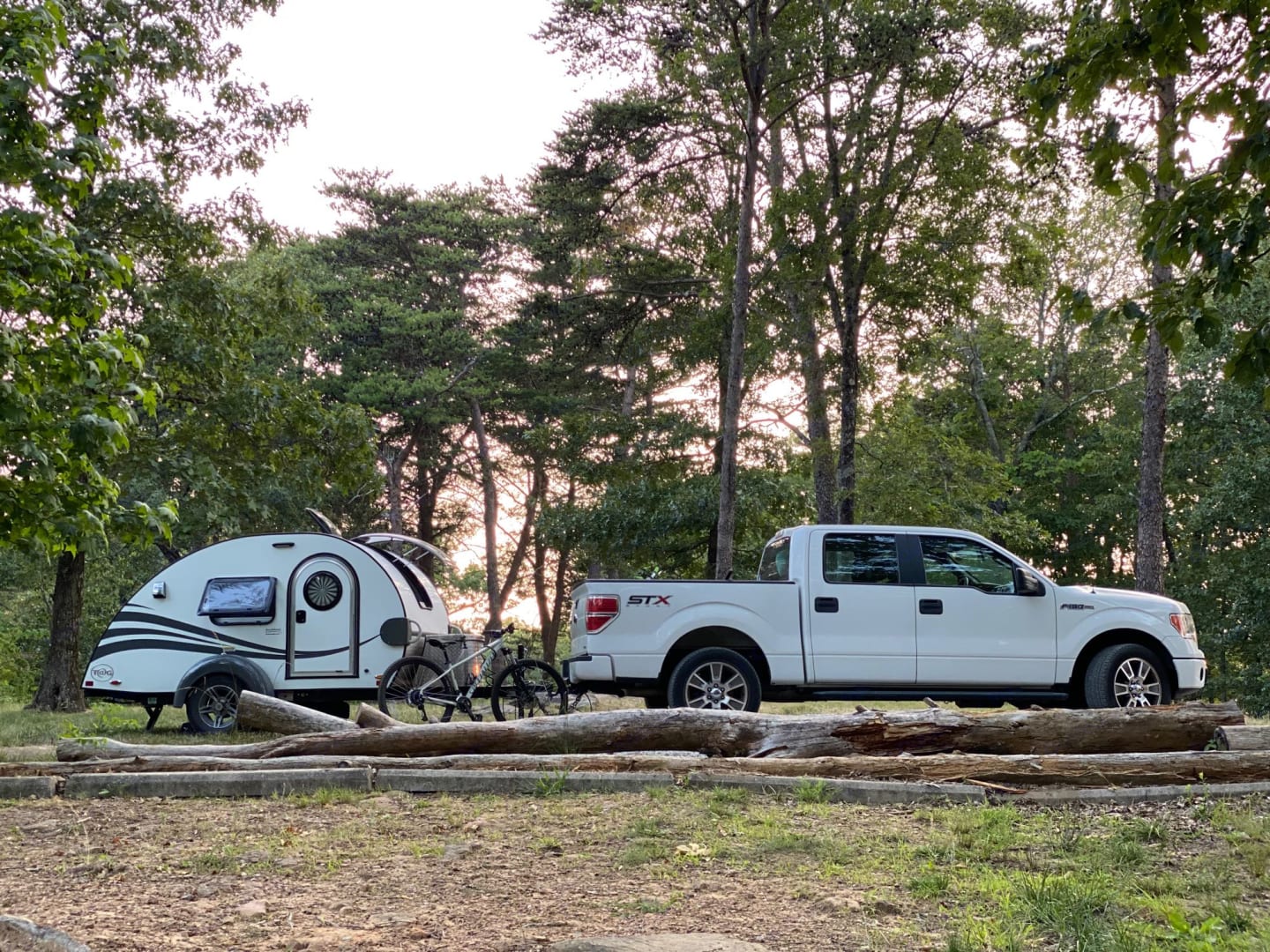 Scenic rv rental setup: white pickup truck towing a teardrop camper with bikes at a wooded campsite.|This white teardrop RV rental