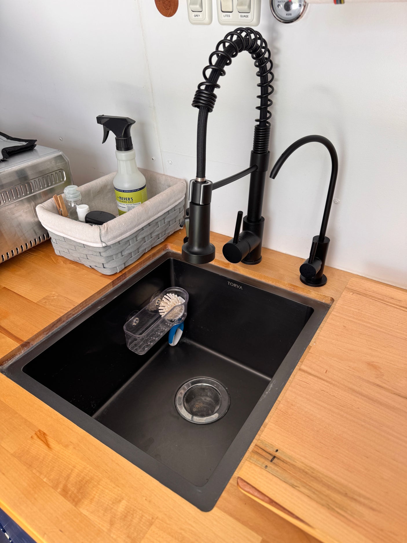 Sleek RV rental kitchen featuring a black sink, dual modern faucets, wood counter, and organized cleaning supplies.