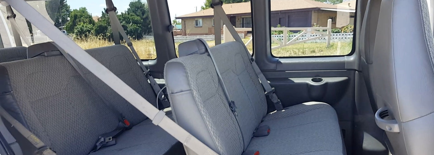 Inside an RV rental van: multiple rows of gray fabric seats, with seatbelts, looking out at houses.