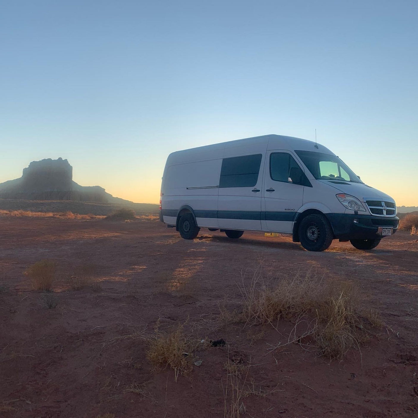 An RV rental, a white camper van, is parked at sunset in a vast desert with a distant mesa. Adventure awaits!