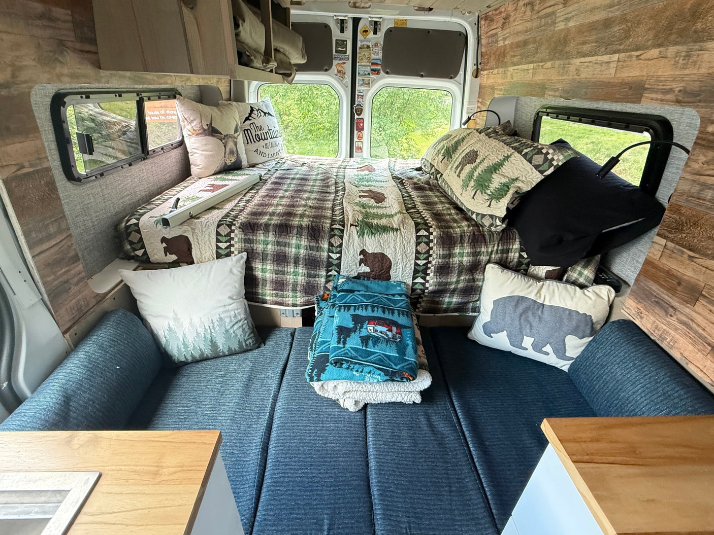 Rustic RV rental interior featuring a cozy bed with plaid bedding, bear & mountain pillows, and bench seating.