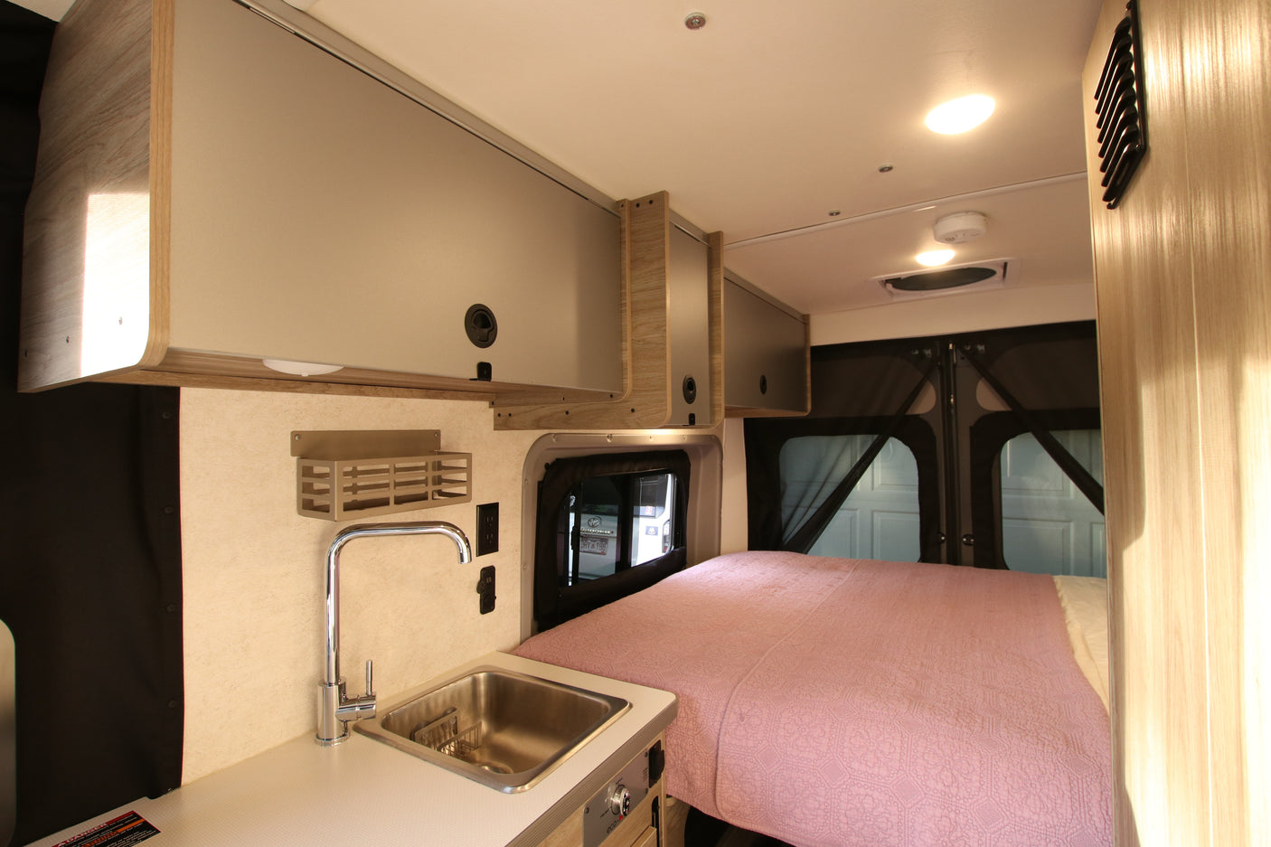 Cozy RV rental interior with a kitchen sink, a bed covered in a pink blanket, and light wood/tan overhead cabinets.