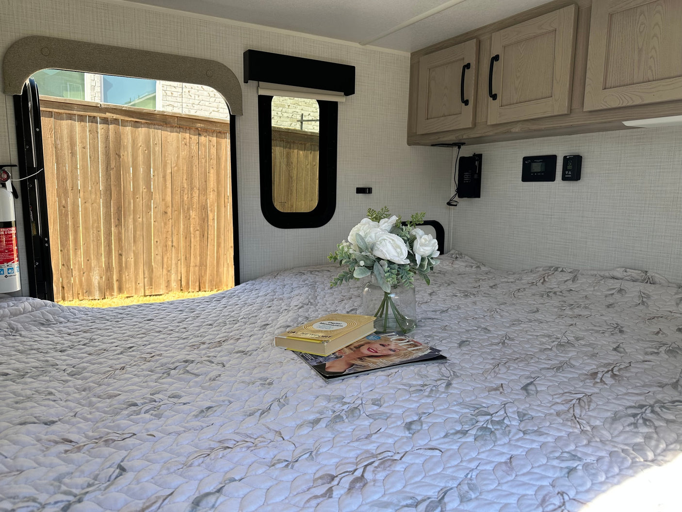 RV rental: Bed with leafy quilt, sunlit windows looking at a fence, overhead cabinets, and a vase of white flowers.