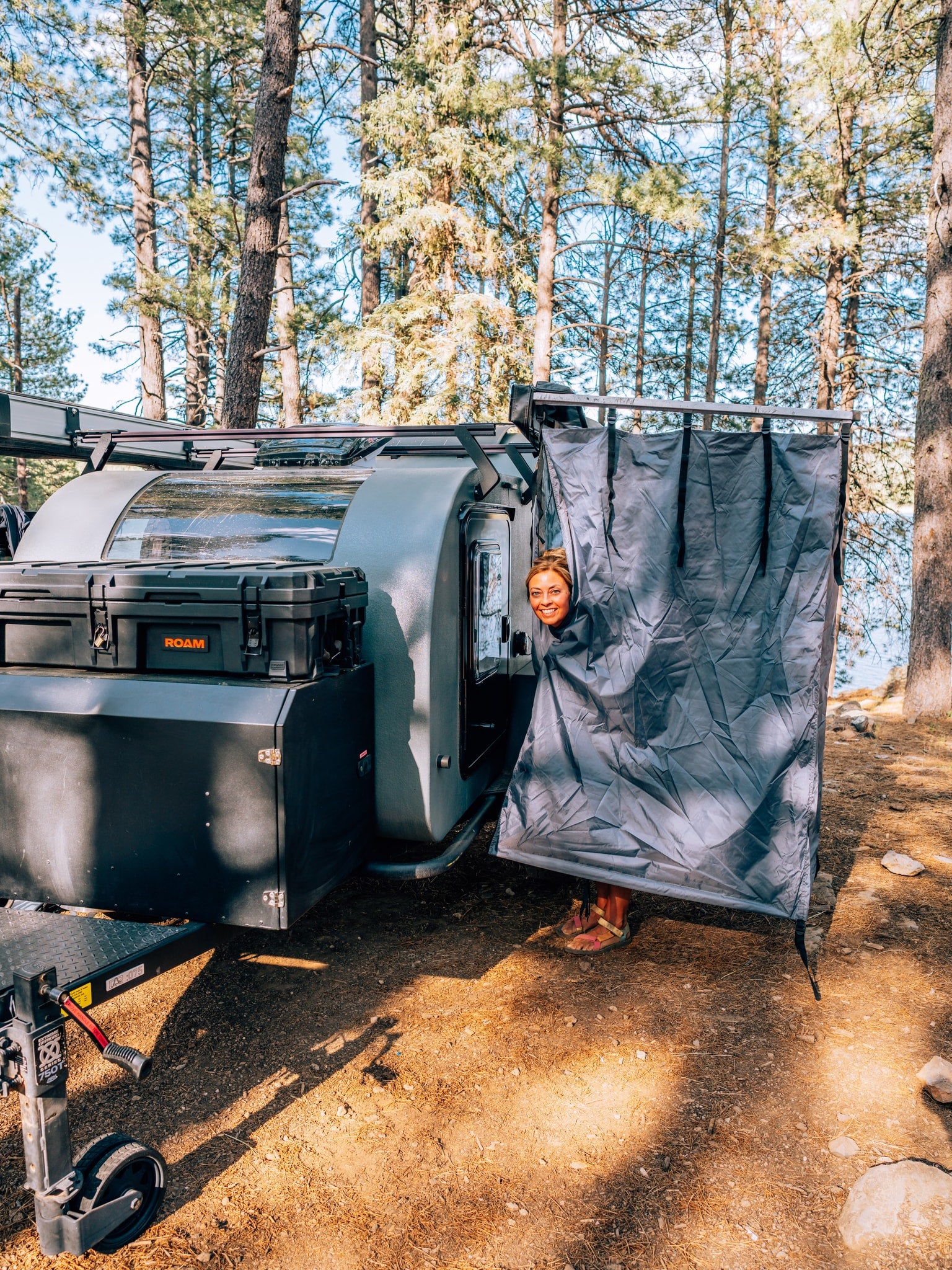 A smiling woman emerges from an outdoor shower attached to her adventure rv rental, set against a forest and lake.