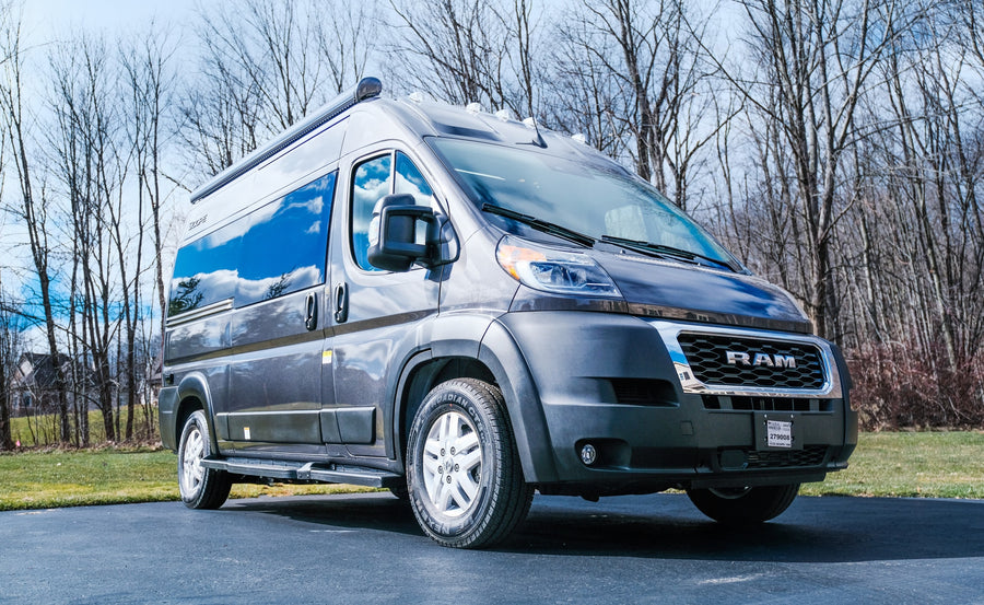 Experience adventure! This gray Ram ProMaster RV rental van is ready for your next trip, parked on an asphalt driveway with bare trees in the background.