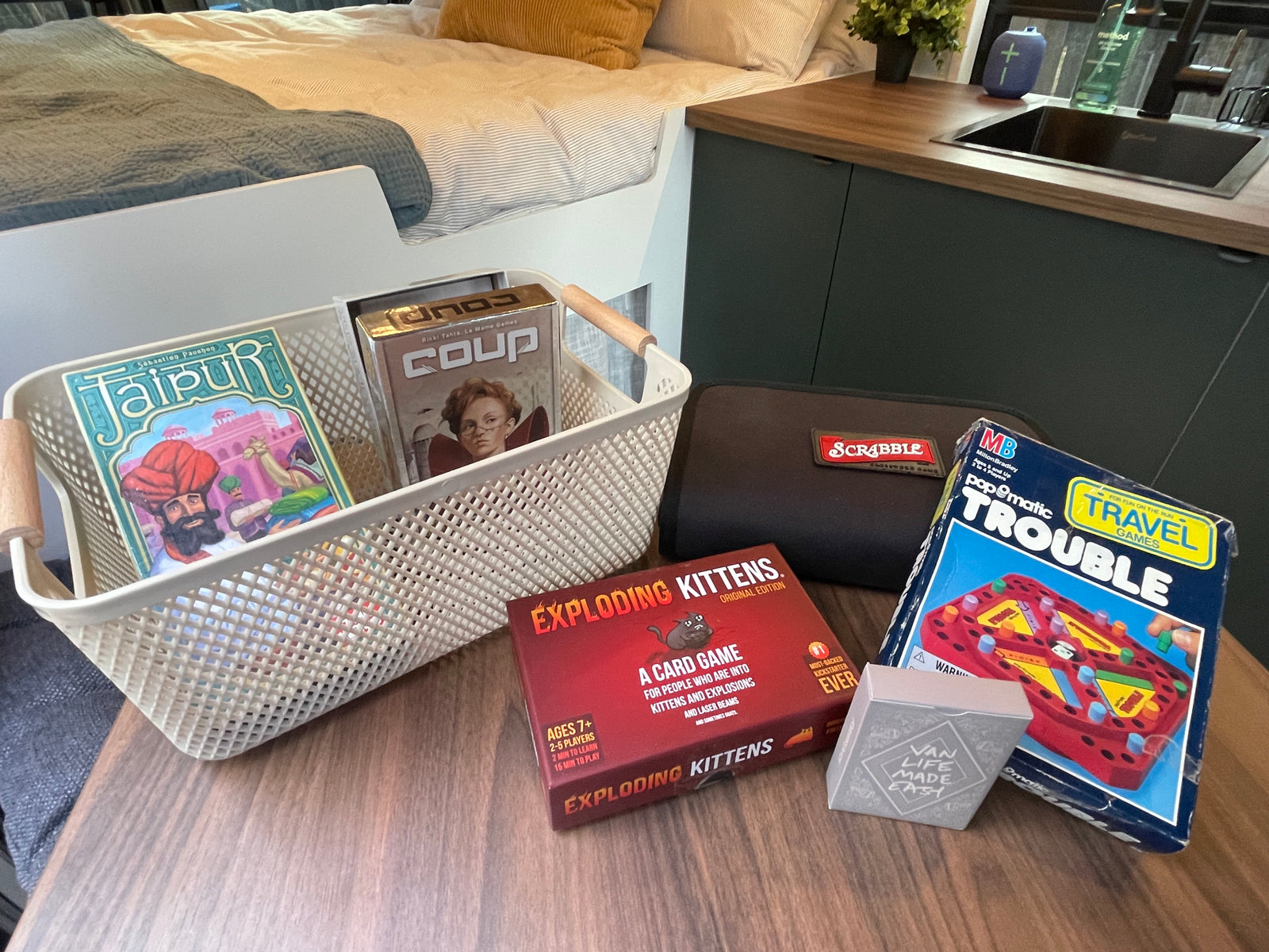 An RV rental's cozy setup: a basket of games (Jaipur, Coup), Exploding Kittens, Scrabble, and Travel Trouble, next to a bed & kitchenette.