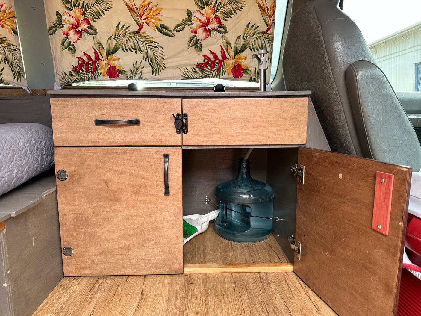 RV rental: Inside a campervan, a custom wood kitchenette with sink, water jug, storage, and floral curtains.
