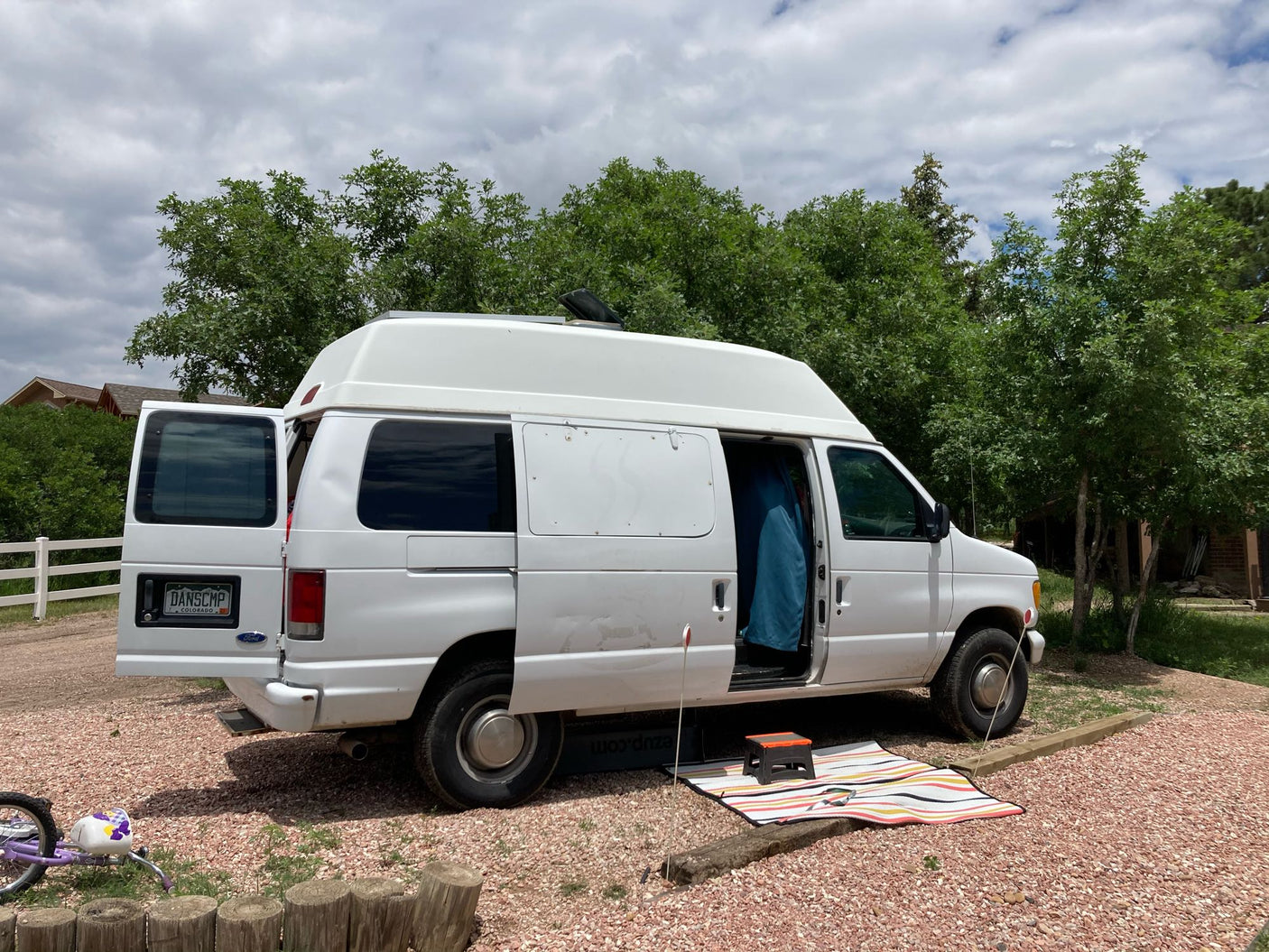 White high-top Ford camper van, ideal for rv rental, parked on gravel with open side/rear doors and outdoor setup.
