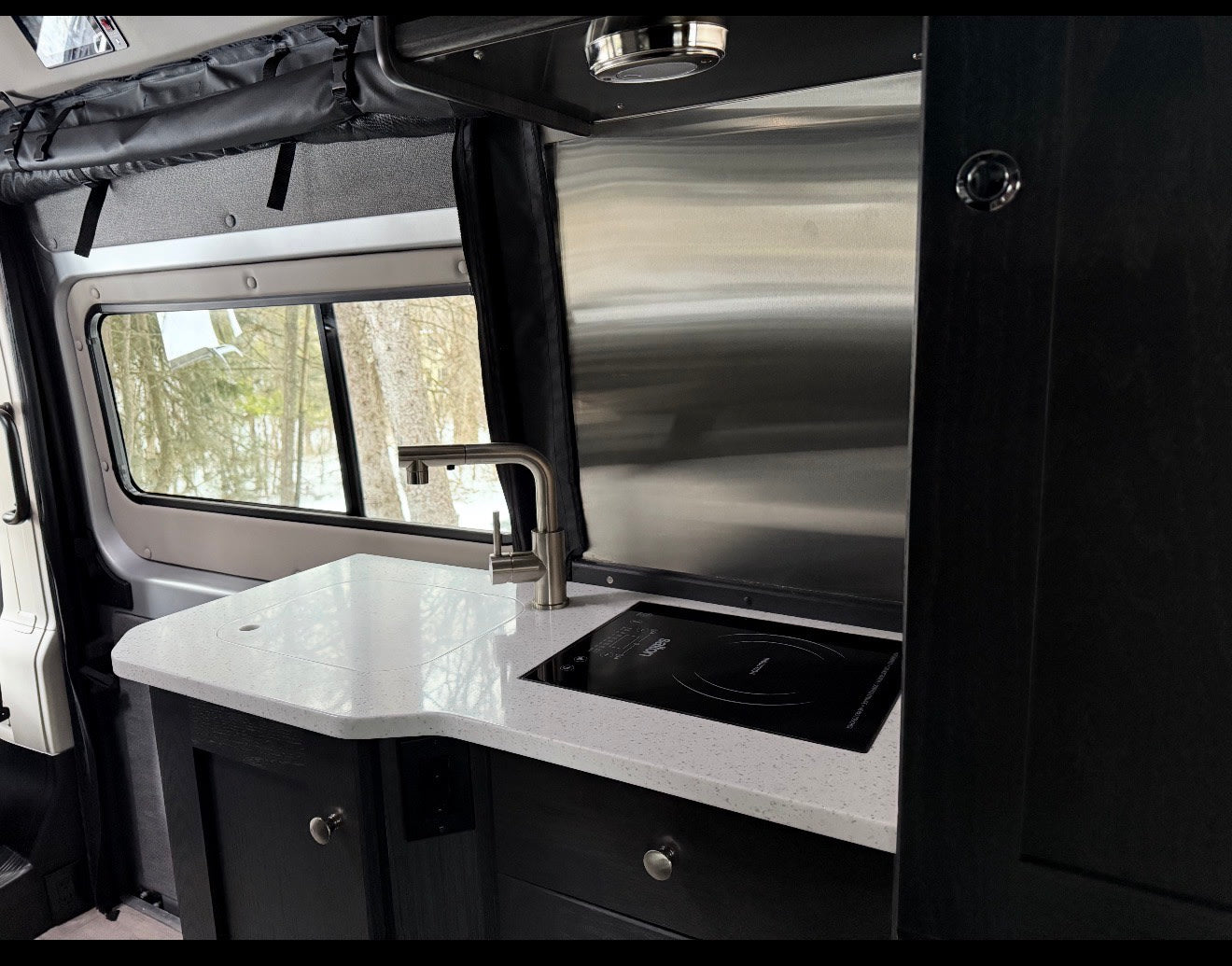 Modern RV rental kitchen with white speckled countertop, induction cooktop, and faucet, featuring a snowy view. — photo 10