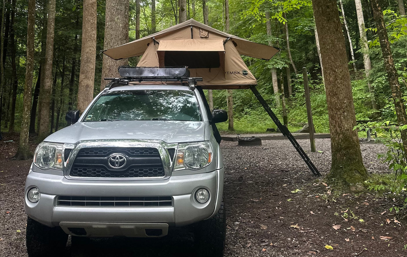 Enjoy nature! This truck with a rooftop tent offers a unique alternative to an RV rental for camping in the woods.|A silver pickup truck with a TMBK rooftop tent set up for camping in a lush forest campsite