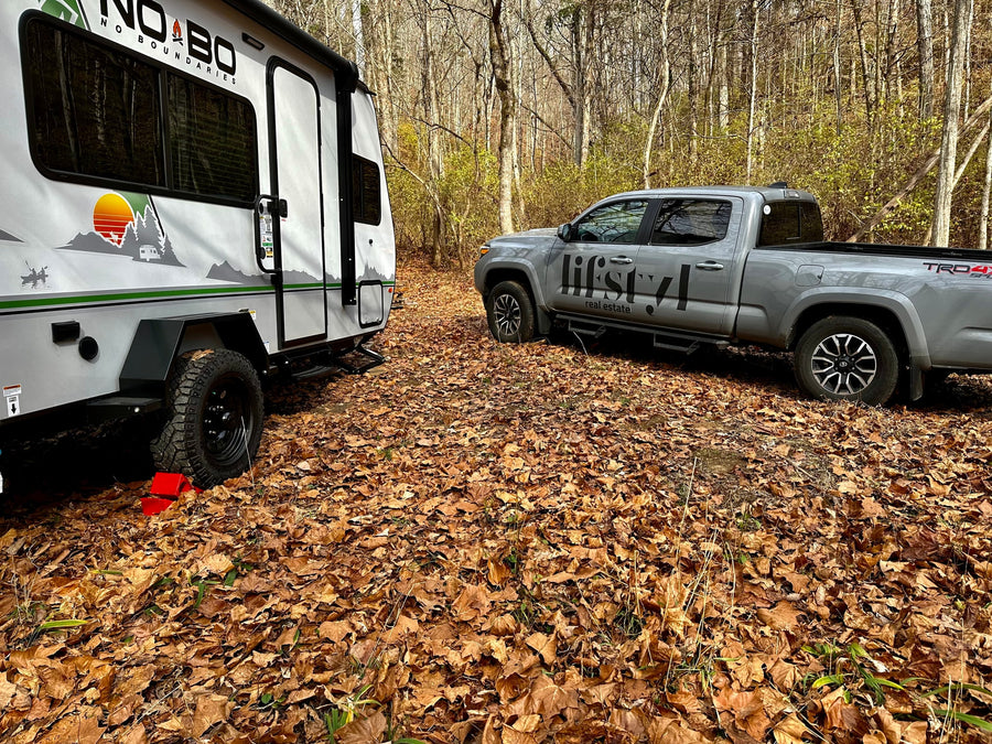 A grey pickup truck and white travel trailer ('No Boundaries' RV) parked in a leaf-covered forest campsite, ideal for rv rental.