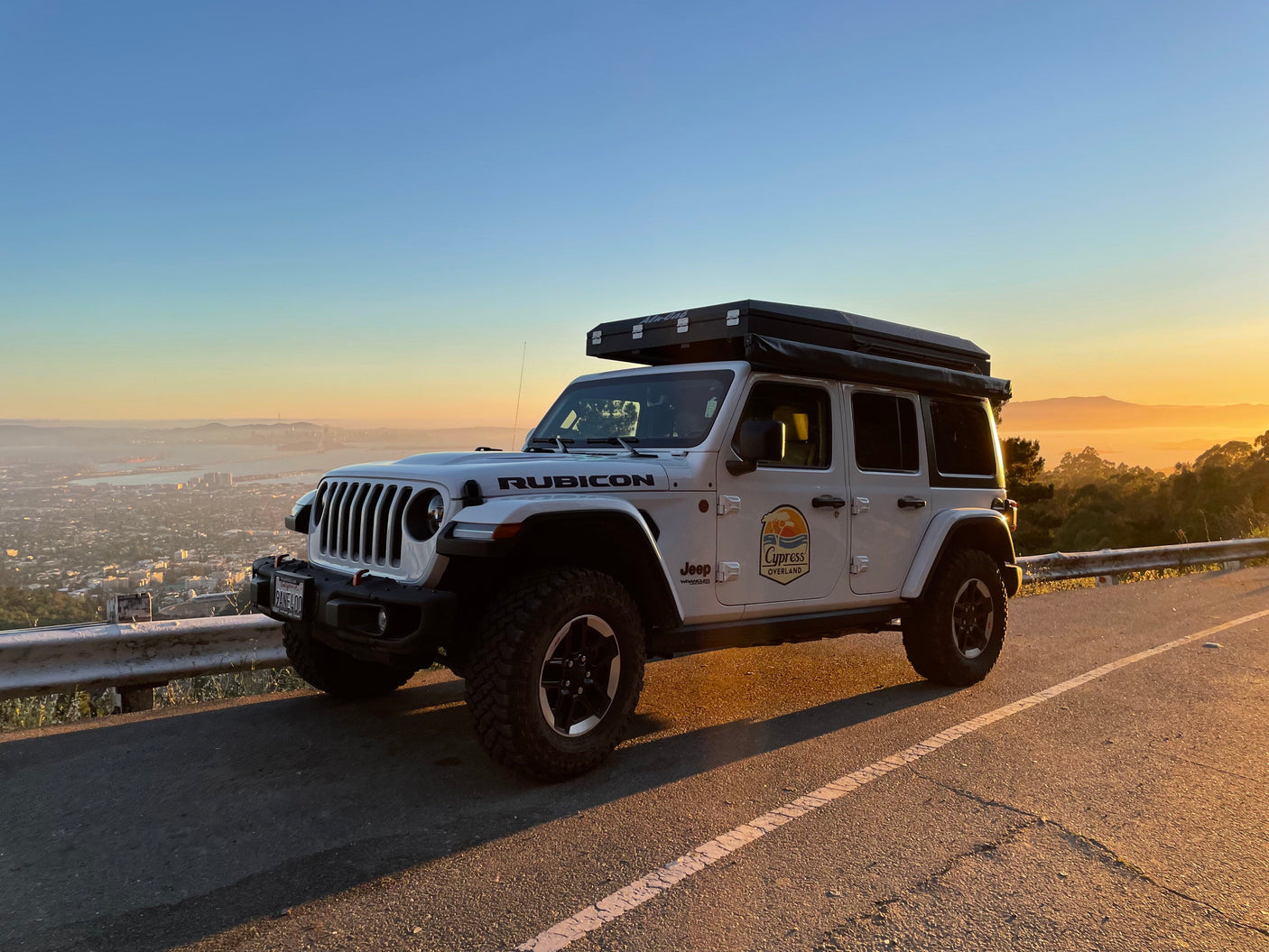 White Jeep Rubicon with rooftop tent, perfect for an overland RV rental adventure, overlooking a hazy city at sunset.