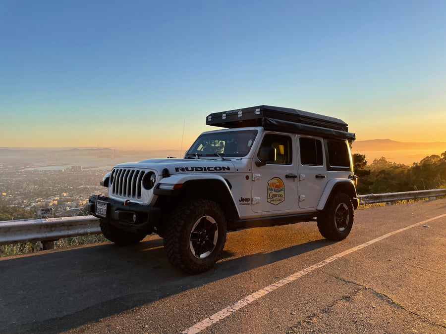 White Jeep Rubicon with rooftop tent, perfect for an overland RV rental adventure, overlooking a hazy city at sunset.