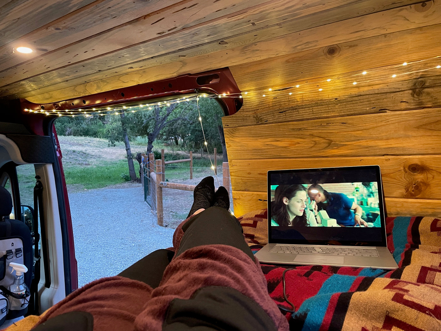 Cozy RV rental: Person relaxing inside a wood-paneled van, watching a movie on a laptop with fairy lights & nature view.