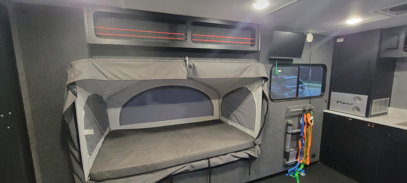 RV rental interior: grey fold-down bunk, overhead storage with red bungees, TV, and compact kitchen with 'Flyer' appliance.