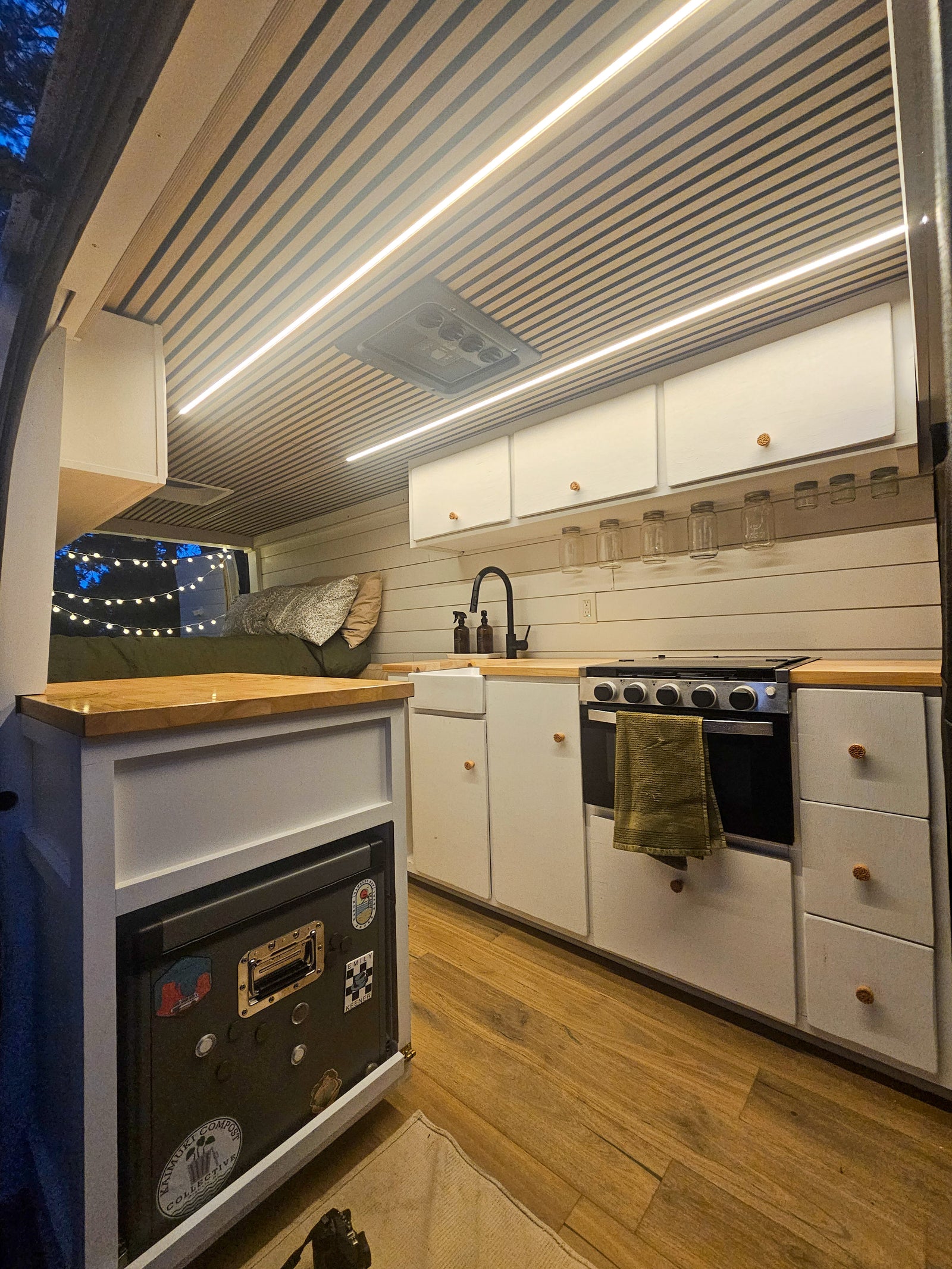 Inside a modern RV rental: a compact kitchen with a stove, sink, and cabinets, plus a cozy sleeping area, all brightly lit.