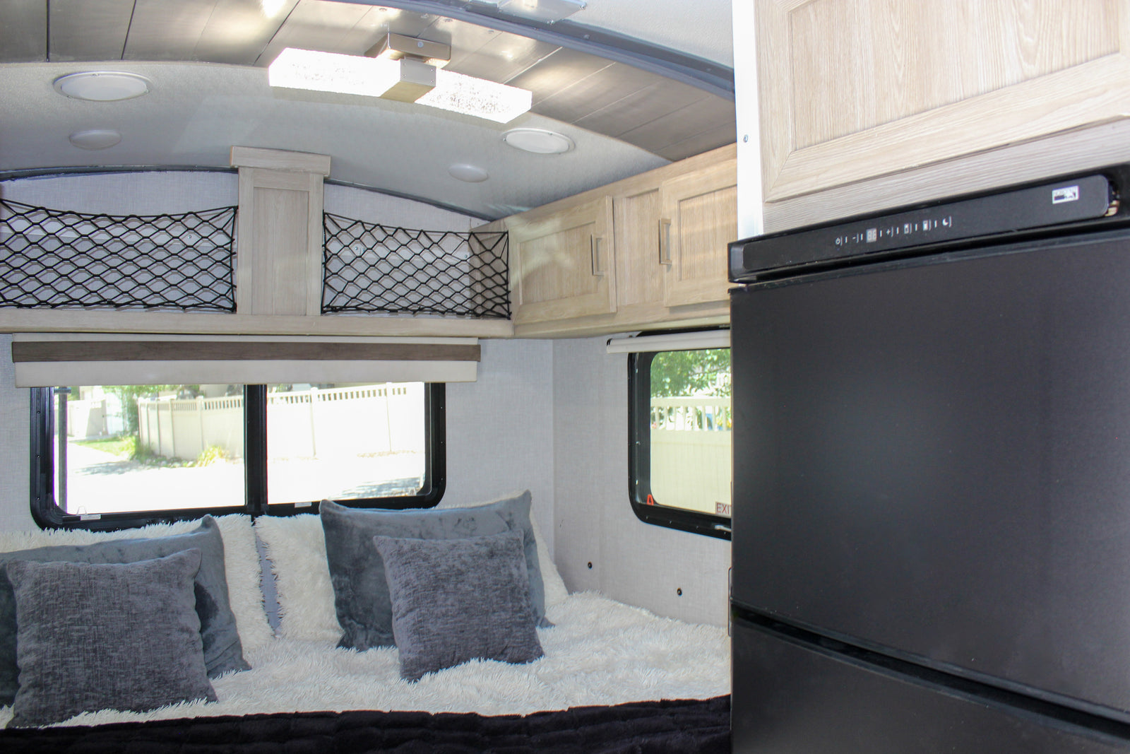 Modern RV rental interior featuring a bed with plush pillows, overhead cabinets, and a compact fridge.